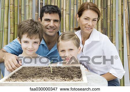 Stock Photo - Family together, smiling at camera, potted seedling in foreground. Fotosearch