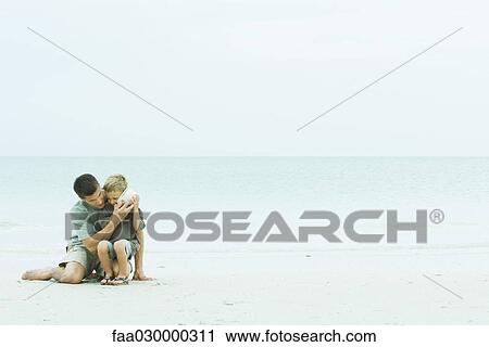 Stock Image - Father and son at the beach, man holding seashell up to boy's ear. Fotosearch