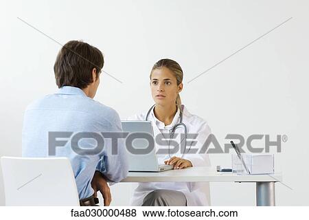 Stock Photo - Female doctor sitting across from male patient at desk, using laptop computer. Fotosearch