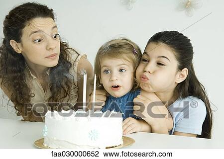 Stock Image - Girl helping younger sister blow out candles on birthday cake, mother watching. Fotosearch