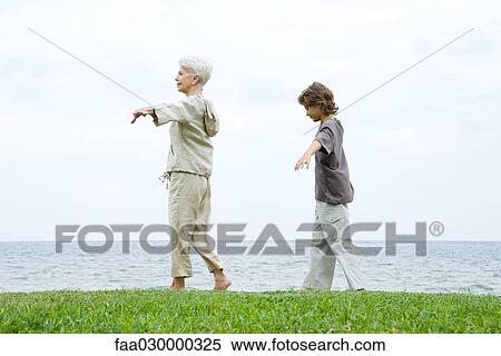 Stock Photography - Grandmother and grandson walking at seashore, arms out, side view. Fotosearch