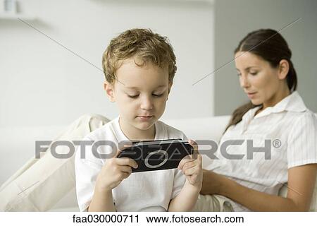 Little boy playing handheld video game, his mother sitting on sofa behind him View Large Photo Image Stock Image - Little boy playing handheld video game, his mother sitting on sofa behind him. Fotosearch