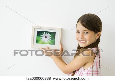 Stock Photo - Little girl holding framed picture against wall, smiling over shoulder at camera. Fotosearch