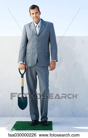 Man in suit, standing on small patch of artificial turf, holding shovel, looking at camera View Large Photo Image Stock Photograph - Man in suit, standing on small patch of artificial turf, holding shovel, looking at camera. Fotosearch