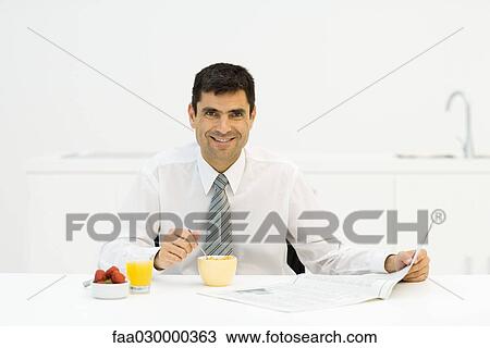 Stock Image - Man sitting at breakfast table with newspaper, smiling at camera. Fotosearch