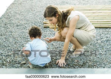 Stock Image - Mother and toddler crouching on gravel, looking at rocks. Fotosearch