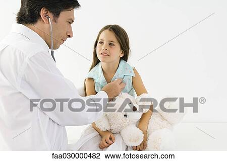 Stock Image - Pediatrician listening to girl's heart with stethoscope, girl holding teddy bear. Fotosearch