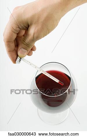 Stock Image - Person holding thermometer over glass of red wine, cropped view of hand. Fotosearch
