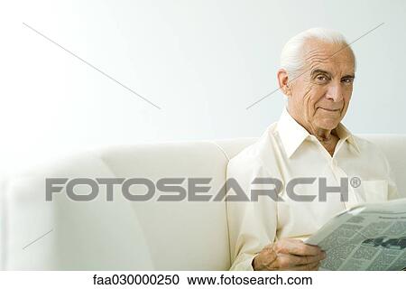 Senior man sitting on sofa, holding newspaper, smiling at camera View Large Photo Image Stock Image - Senior man sitting on sofa, holding newspaper, smiling at camera. Fotosearch