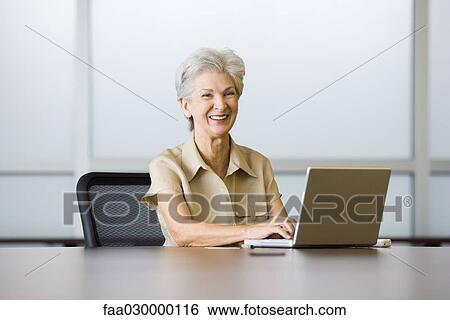 Senior woman sitting at desk, using laptop, laughing View Large Photo Image Stock Photograph - Senior woman sitting at desk, using laptop, laughing. Fotosearch