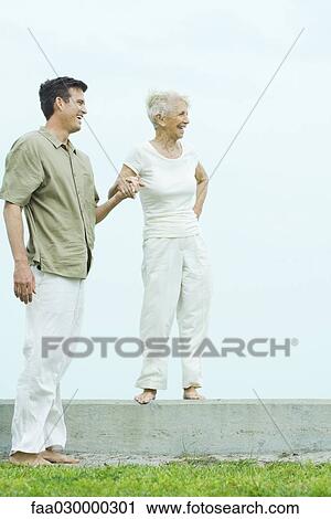 Senior woman standing on ledge, holding adult son's hand, both looking away, smiling View Large Photo Image Stock Image - Senior woman standing on ledge, holding adult son's hand, both looking away, smiling. Fotosearch