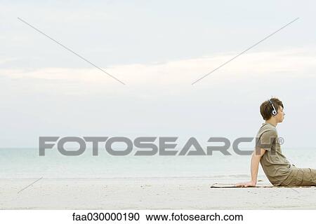 Stock Image - Teen boy sitting on beach listening to headphones, cropped. Fotosearch
