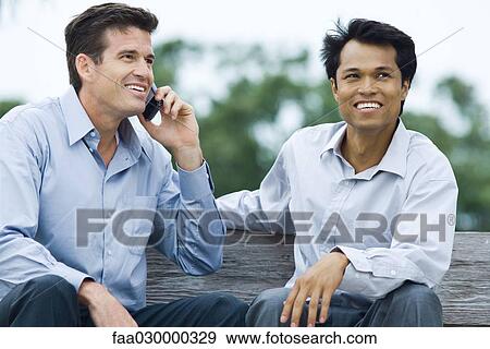 Two men sitting on bench, smiling, one using cell phone View Large Photo Image Stock Photo - Two men sitting on bench, smiling, one using cell phone. Fotosearch