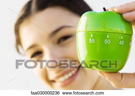 Stock Photograph - Woman holding up kitchen timer, smiling at camera, focus on foreground. Fotosearch