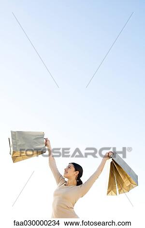 Picture - Woman holding up shopping bags, smiling, low angle view. Fotosearch