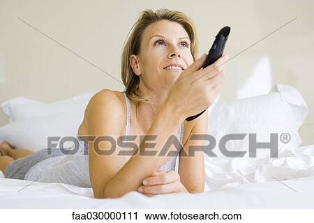 Stock Image - Woman lying on bed, changing channel with remote control. Fotosearch