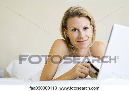 Woman lying on bed, using credit card and laptop, smiling at camera View Large Photo Image Stock Image - Woman lying on bed, using credit card and laptop, smiling at camera. Fotosearch