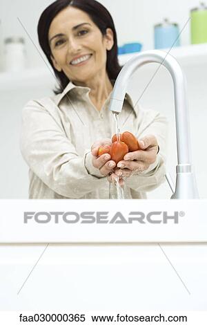 Stock Photography - Woman rinsing roma tomatoes under faucet, smiling at camera. Fotosearch