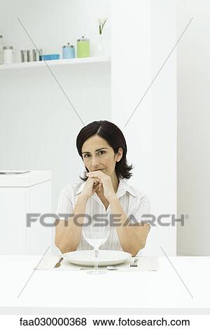 Stock Photo - Woman sitting at dinner table, hands clasped in front of mouth, looking at camera. Fotosearch