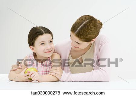 Woman sitting at table with arm around daughter's shoulder, smiling, girl holding apple View Large Photo Image Stock Photo - Woman sitting at table with arm around daughter's shoulder, smiling, girl holding apple. Fotosearch