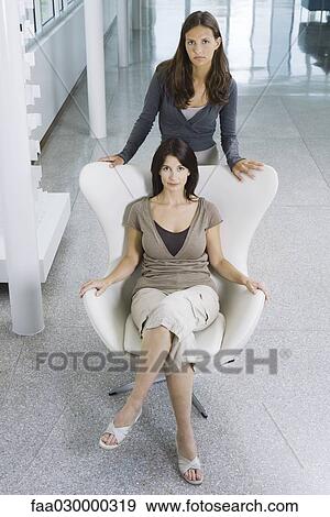 Stock Photo - Woman sitting in chair, teen daughter standing behind her, both looking up at camera. Fotosearch