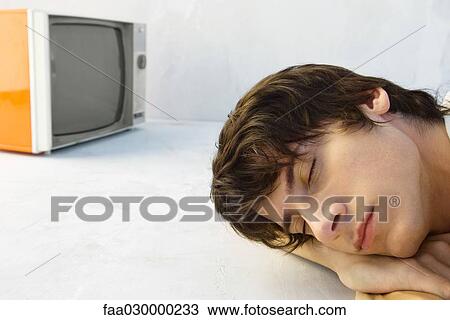 Stock Image - Young man resting head on arms, eyes closed, television in background. Fotosearch