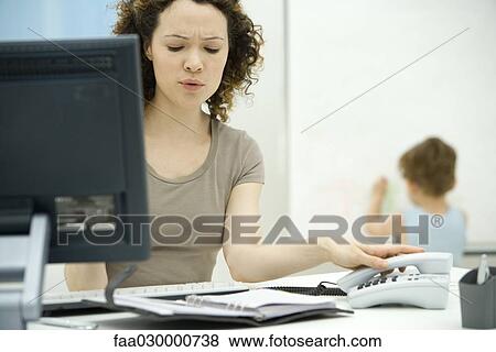 Stock Photo - Young woman hanging up phone and scowling, son drawing on wall in background. Fotosearch