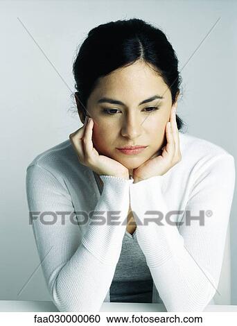 Stock Image - Young woman holding head in hands, looking down, portrait. Fotosearch