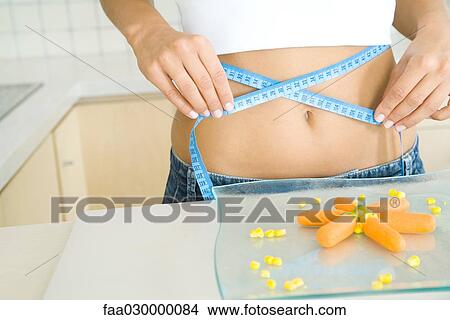 Picture - Young woman holding measuring tape around bare abdomen, plate of veggies in the foreground. Fotosearch