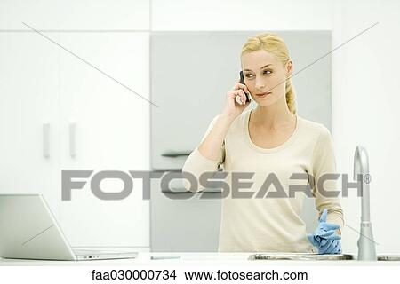 Picture - Young woman standing in kitchen, holding rubber gloves, using cell phone. Fotosearch