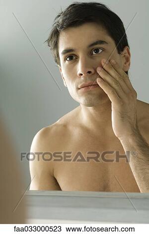Stock Image - Bare-chested man looking at self in mirror, touching face. Fotosearch