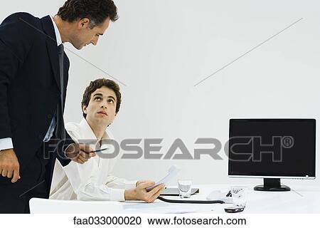 Stock Image - Boss looking over employee's shoulder, employee holding hand over mouthpiece of phone. Fotosearch