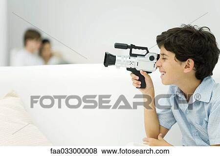 Boy looking through video camera, couple in background, side view View Large Photo Image Stock Photo - Boy looking through video camera, couple in background, side view. Fotosearch