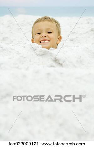 Stock Image - Boy's head emerging from sand, smiling at camera. Fotosearch