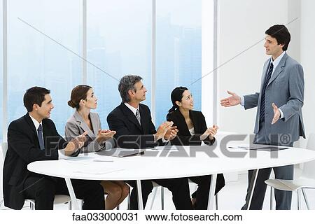 Stock Image - Business people applauding associate's presentation, sitting around table. Fotosearch