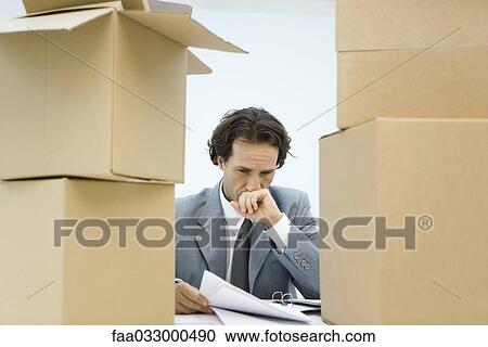 Stock Image - Businessman between stacks of cardboard boxes, studying document, hand over mouth. Fotosearch