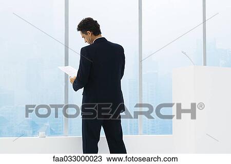 Stock Image - Businessman reviewing document, standing, rear view. Fotosearch