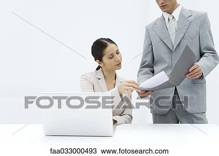 Stock Image - Businessman showing document in folder to female colleague sitting behind laptop, cropped view. Fotosearch