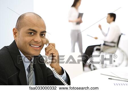 Stock Image - Businessman using phone, smiling at camera, colleagues in background. Fotosearch