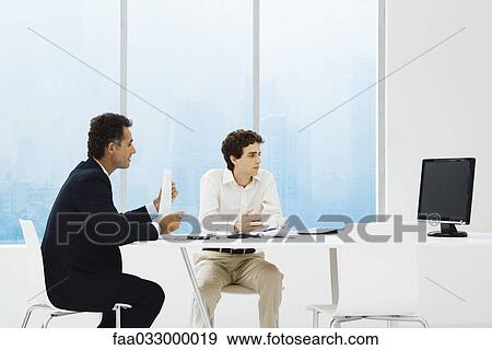 Stock Photo - Businessmen sitting at table, looking at monitor, teleconferencing . Fotosearch