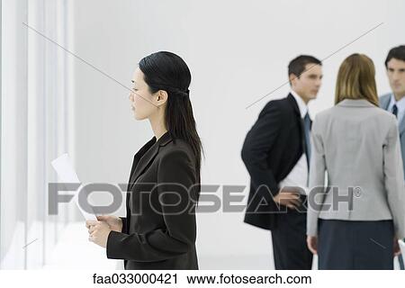 Stock Image - Businesswoman looking out window, holding document, colleagues talking in background. Fotosearch