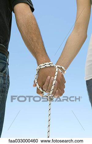 Stock Image - Couple holding hands, bound with rope, cropped view. Fotosearch