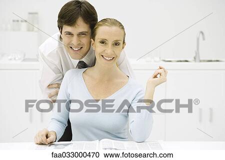 Stock Image - Couple in kitchen, smiling at camera, man leaning over woman's shoulder. Fotosearch