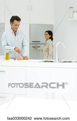 Stock Image - Couple standing in kitchen, man slicing tomato, woman opening refrigerator, both smiling. Fotosearch