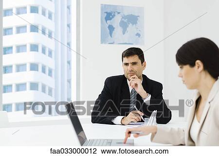 Stock Image - Executive sitting in office with hand under chin, female colleague using laptop in foreground. Fotosearch