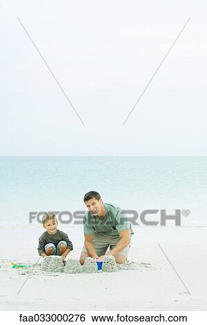 Stock Photograph - Father and son making sand castles at the beach, smiling at camera. Fotosearch