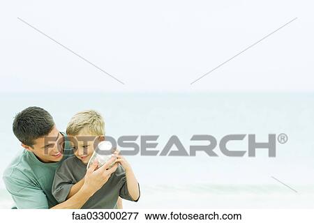 Stock Photo - Father holding seashell up to son's ear at the beach. Fotosearch