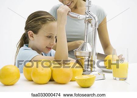 Picture - Girl making fresh orange juice with citrus press, mother in background. Fotosearch