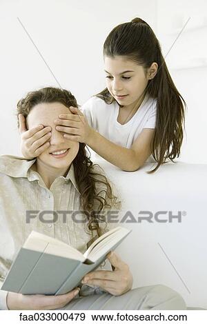 Stock Photo - Girl standing with hands over her mother's eyes, woman holding book. Fotosearch