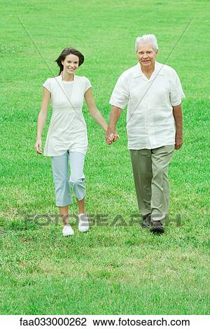 Stock Image - Grandfather and teen granddaughter walking outdoors, holding hands, smiling at camera. Fotosearch